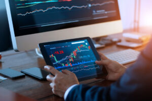 Businessman checking stock market on digital tablet and a desktop computer with stock exchange graph on screen. Financial stock market data analysis